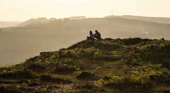 Curbar edge couple This landscape photograph shows a couple sitting on Curbar Edge, which is a prominent gritstone escarpment in the Peak District, Derbyshire, United Kingdom. The scene captures Curbar Edge during a spring evening, with golden sunlight illuminating the rural landscape and casting long shadows over the greenery and rock formations. In the distance, rolling hills and patchwork fields characteristic of the Peak District extend toward the horizon, showcasing the natural beauty of the area. The photograph highlights the tranquil nature of the Derbyshire countryside, with layers of hills and scattered trees under a warm, soft light typical of spring evenings.
